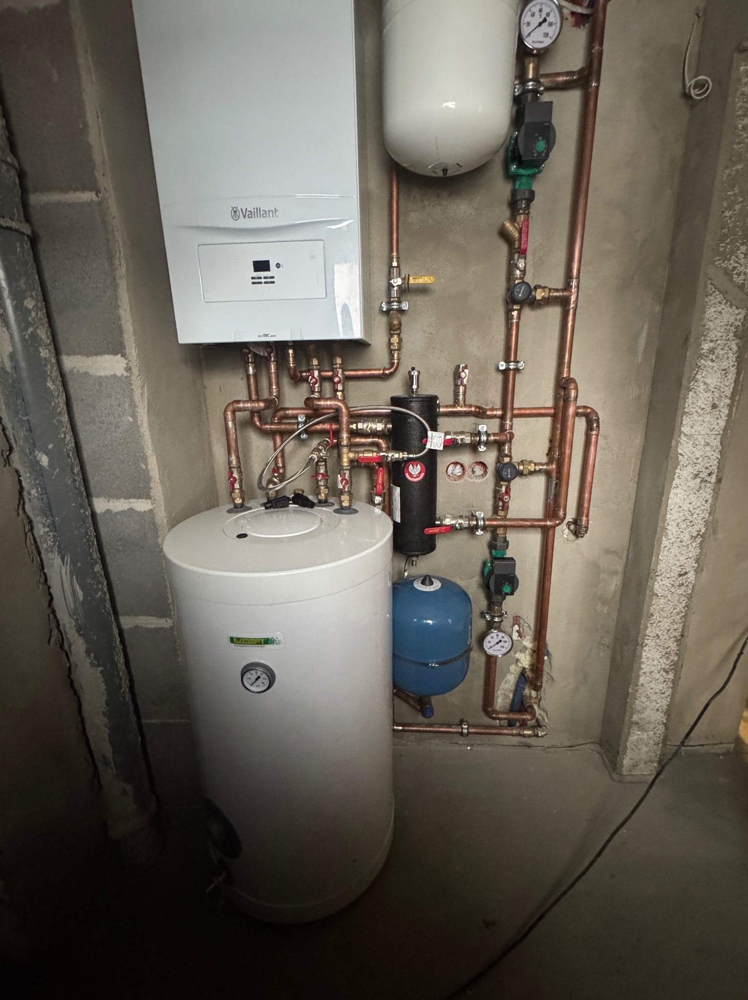 Zmontowana instalacja grzewcza z kotłem Vaillant, zbiornikiem i miedzianymi rurami na surowej ścianie. Widoczne manometry i zawory.
