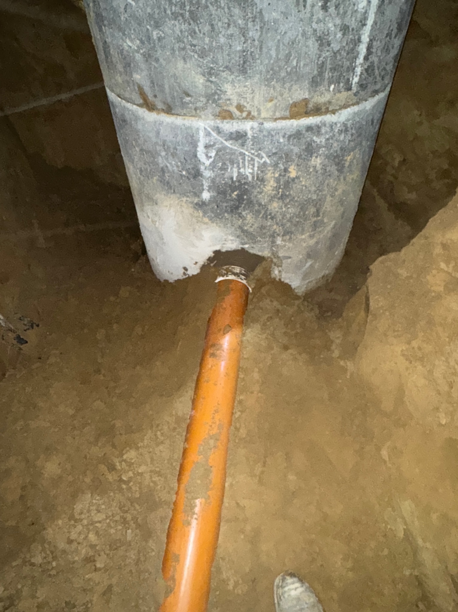 Szara studnia betonowa z podłączoną pomarańczową rurą kanalizacyjną w wykopie ziemnym. Widoczne ślady zaprawy i ziemi na elementach.