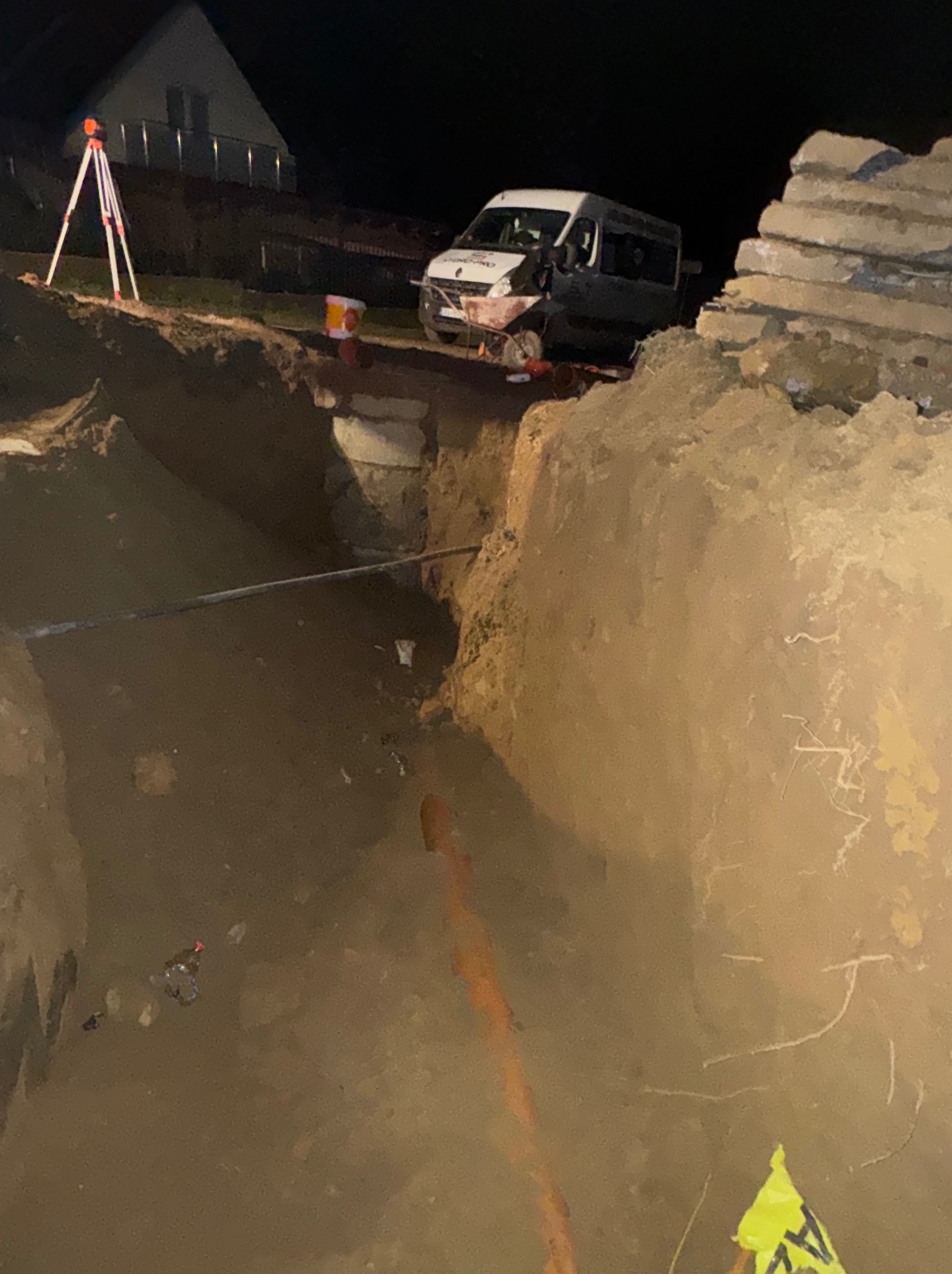 Wykop pod instalację wod-kan nocą, widoczny rów z rurą, samochód dostawczy i niwelator laserowy. Prace ziemne przy oświetleniu.
