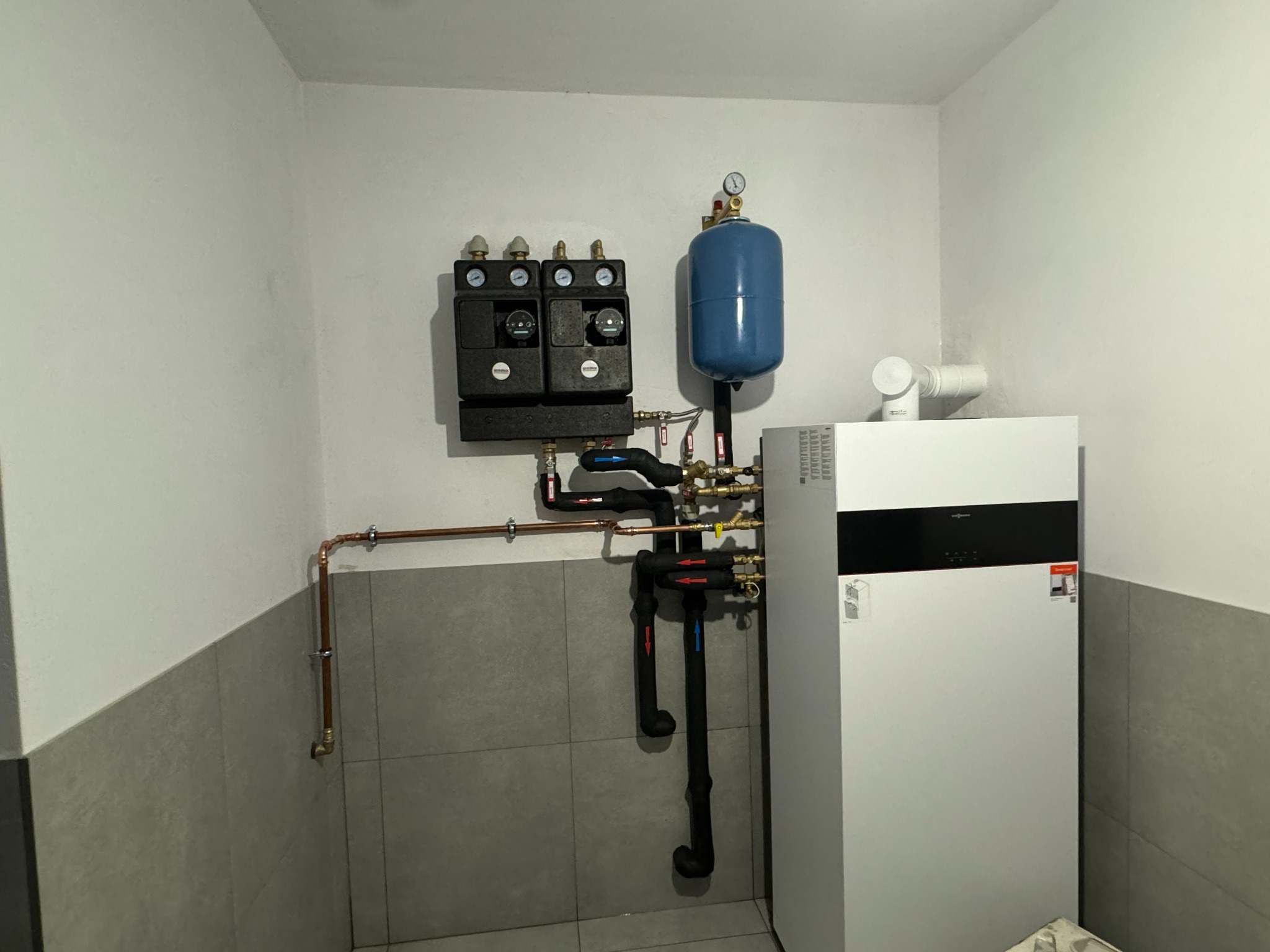 Nowoczesna instalacja grzewcza Viessmann z niebieskim naczyniem wzbiorczym, miedzianymi rurami i czarnymi rozdzielaczami w pomieszczeniu technicznym z szarymi płytkami.