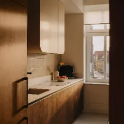 Nowoczesna kuchnia z drewnianymi szafkami w ciepłym odcieniu, jasnym blatem i widokiem na miasto za oknem, oświetlona popołudniowym słońcem.