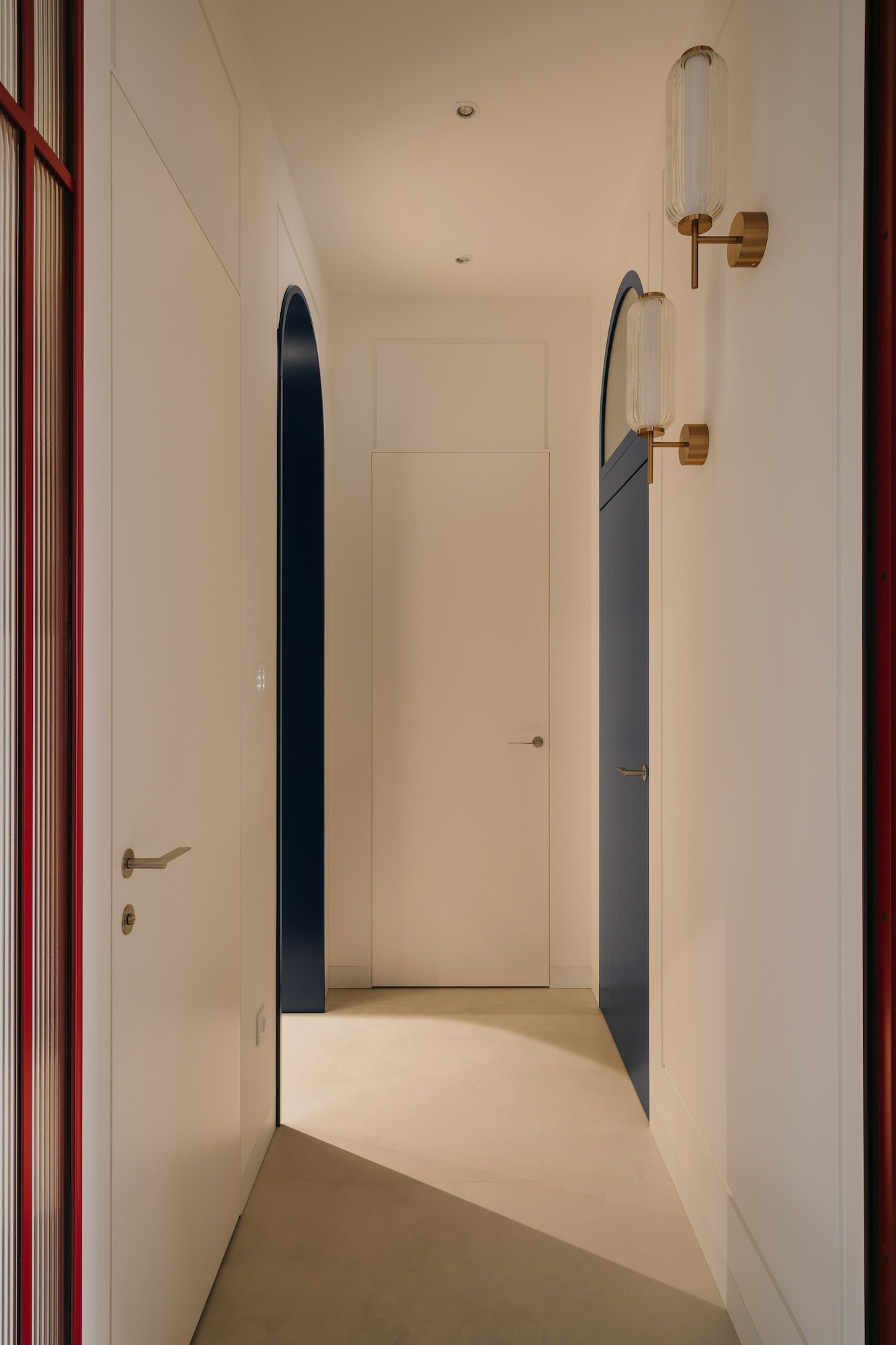 Nowoczesny korytarz z białymi ścianami, ciemnoniebieskimi wnękami drzwiowymi w kształcie łuków, minimalistycznymi lampami ściennymi z ryflowanego szkła i złotymi elementami oraz jasną podłogą...