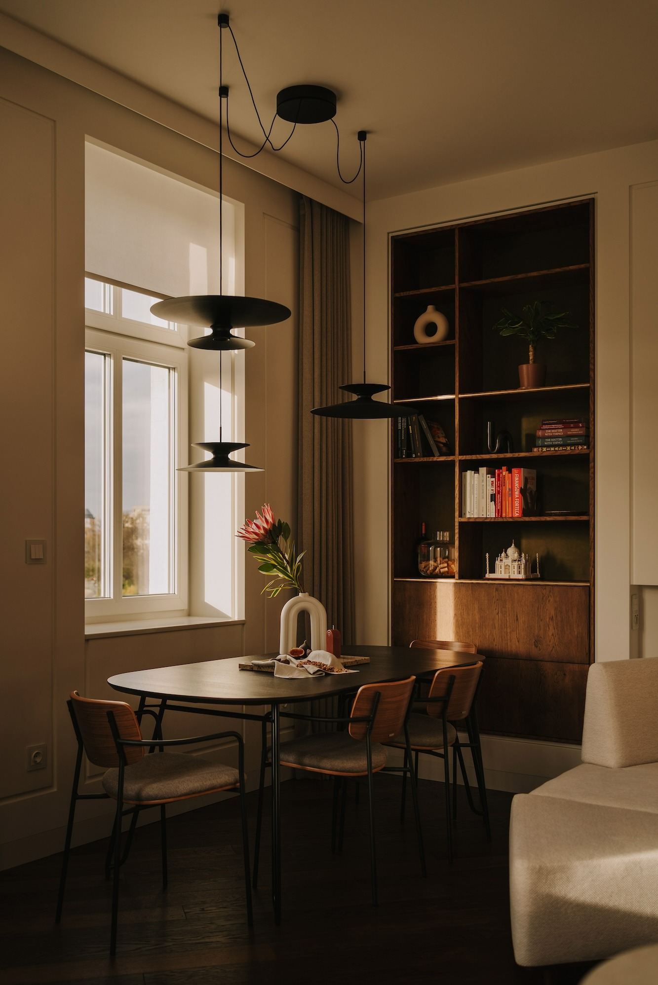 Jadalnia w stylu minimalistycznym z czarnym stołem, drewnianymi krzesłami, trzema okrągłymi lampami wiszącymi i regałem z książkami oraz dekoracjami.