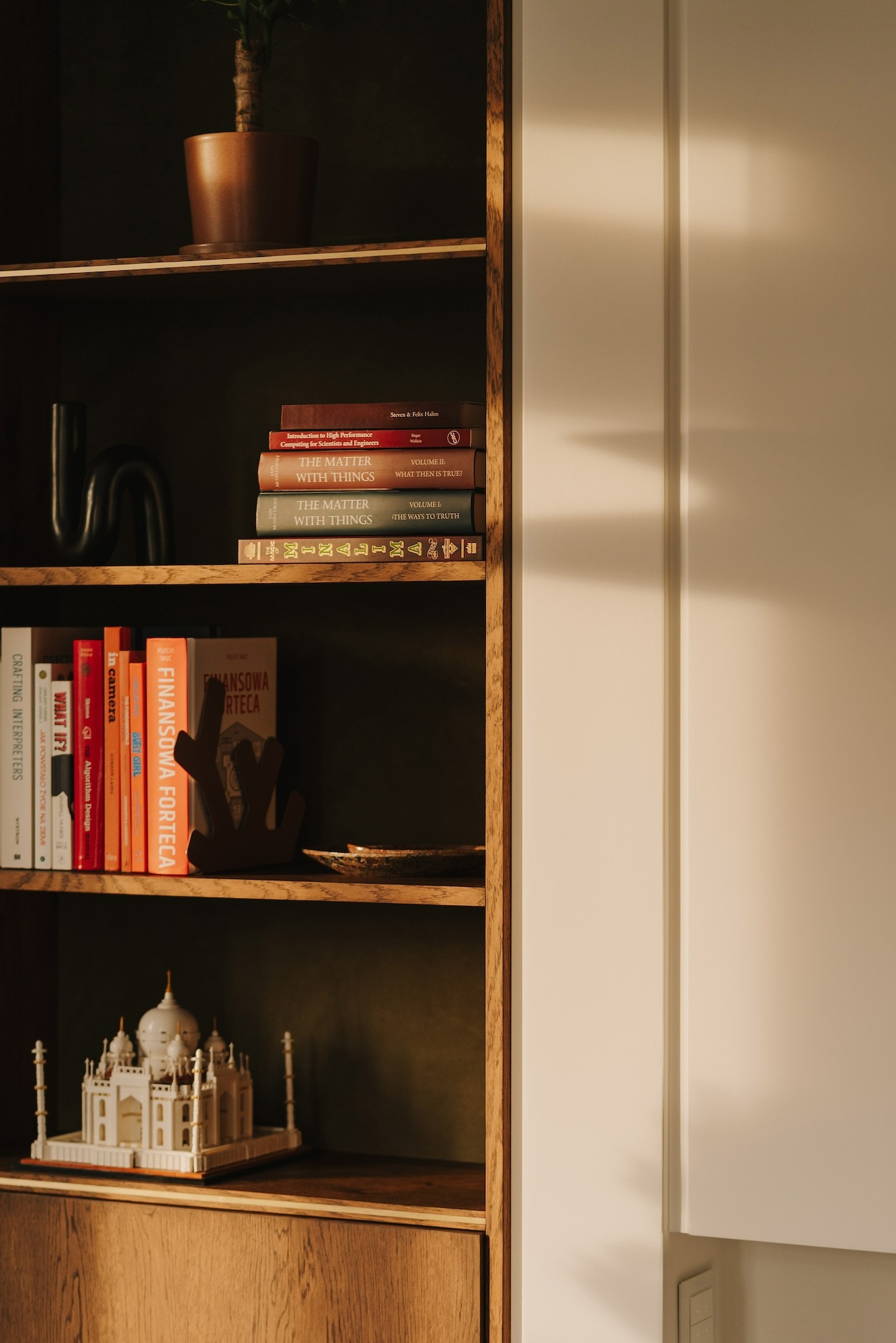 Regał z książkami i dekoracjami, w tym model Taj Mahal, oświetlony ciepłym światłem, z widocznymi detalami okładki książek i teksturą drewna.