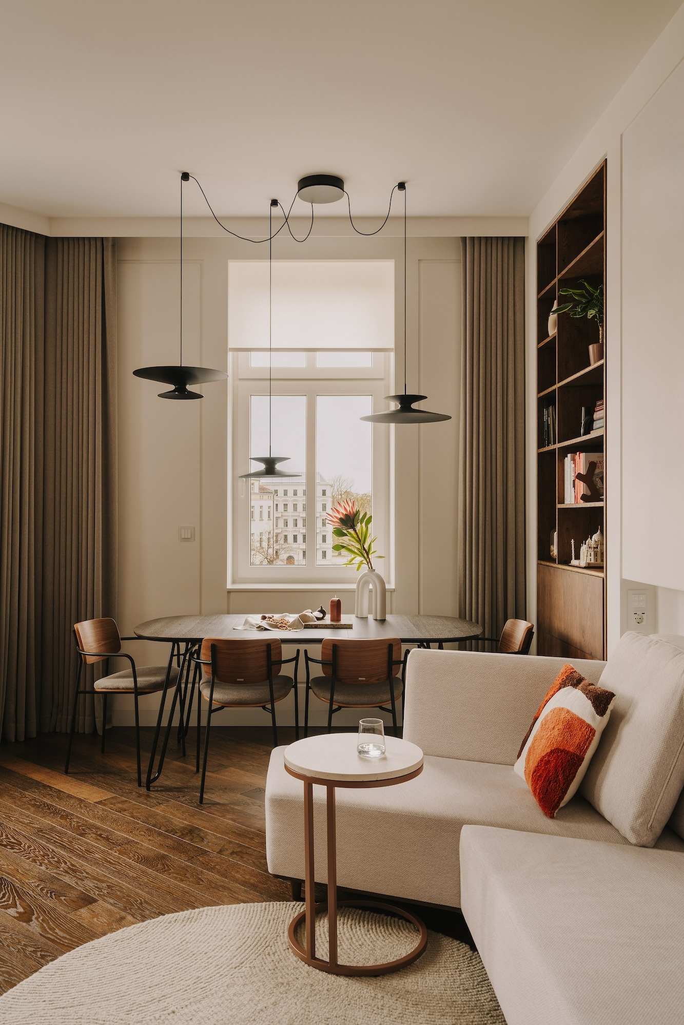 Jasne wnętrze mieszkania z drewnianą podłogą, okrągłym stołem z czarnymi nogami i kwiatem w wazonie na tle okna, minimalistyczna sofa z dekoracyjną poduszką oraz regał na książki.