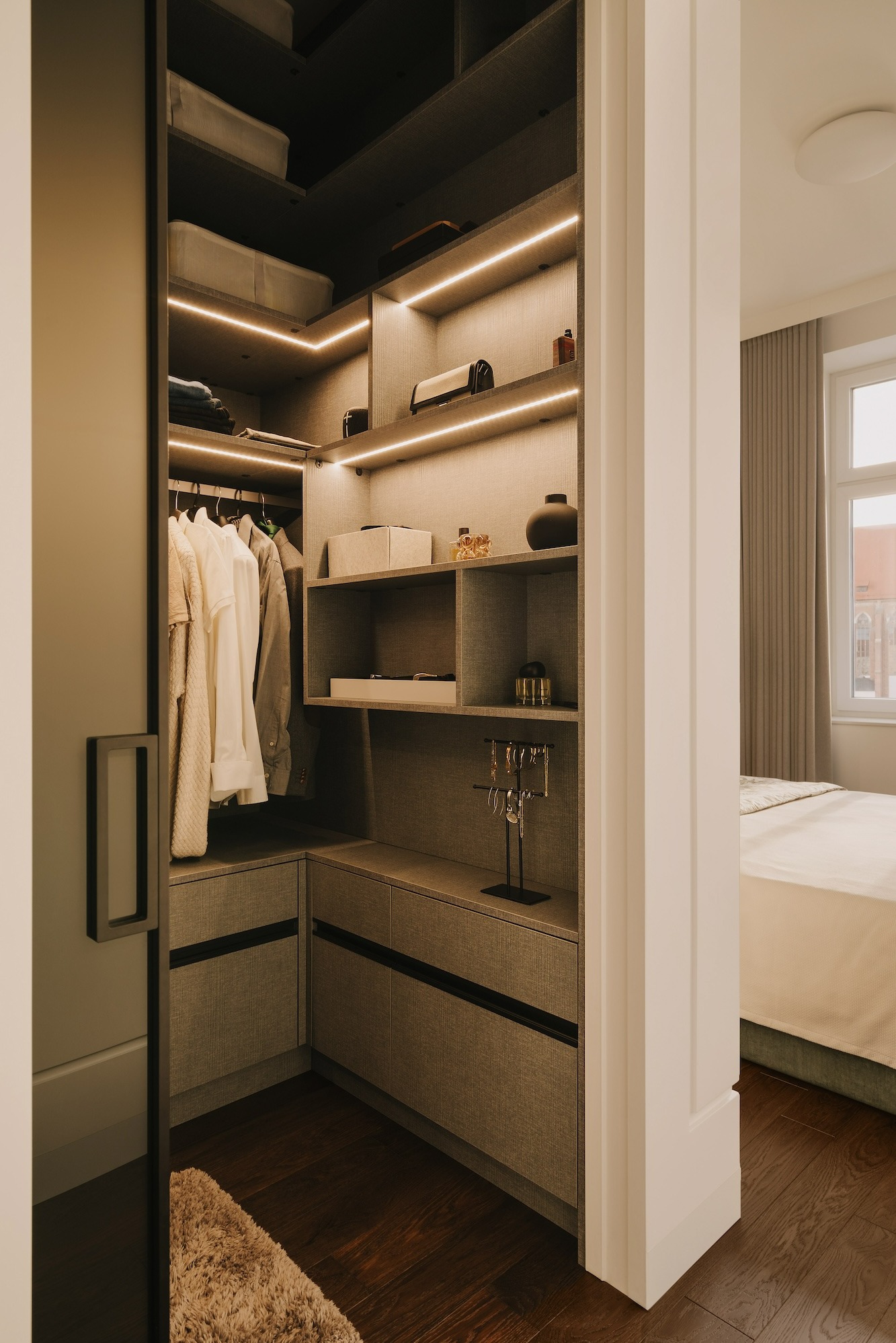 Wnętrze szafy wnękowej z oświetleniem LED, ubrania na wieszakach, pudełka i dekoracje na półkach, widok na sypialnię z łóżkiem i oknem.