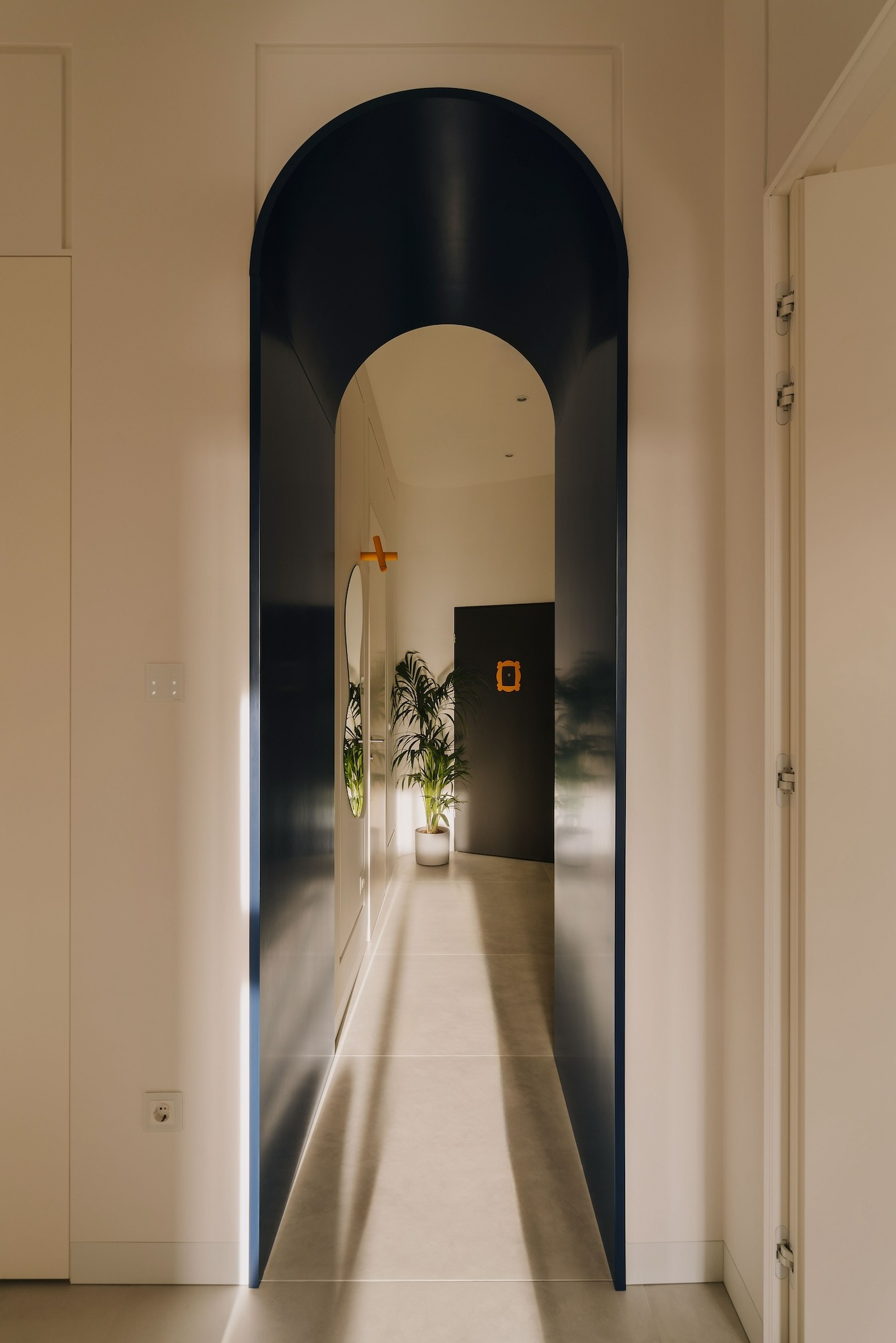Nowoczesny korytarz z ciemnoniebieskim łukowym przejściem, jasnymi ścianami, szarą podłogą i rośliną doniczkową w głębi, z akcentem w postaci pomarańczowego wieszaka i dekoracji na drzwiach.