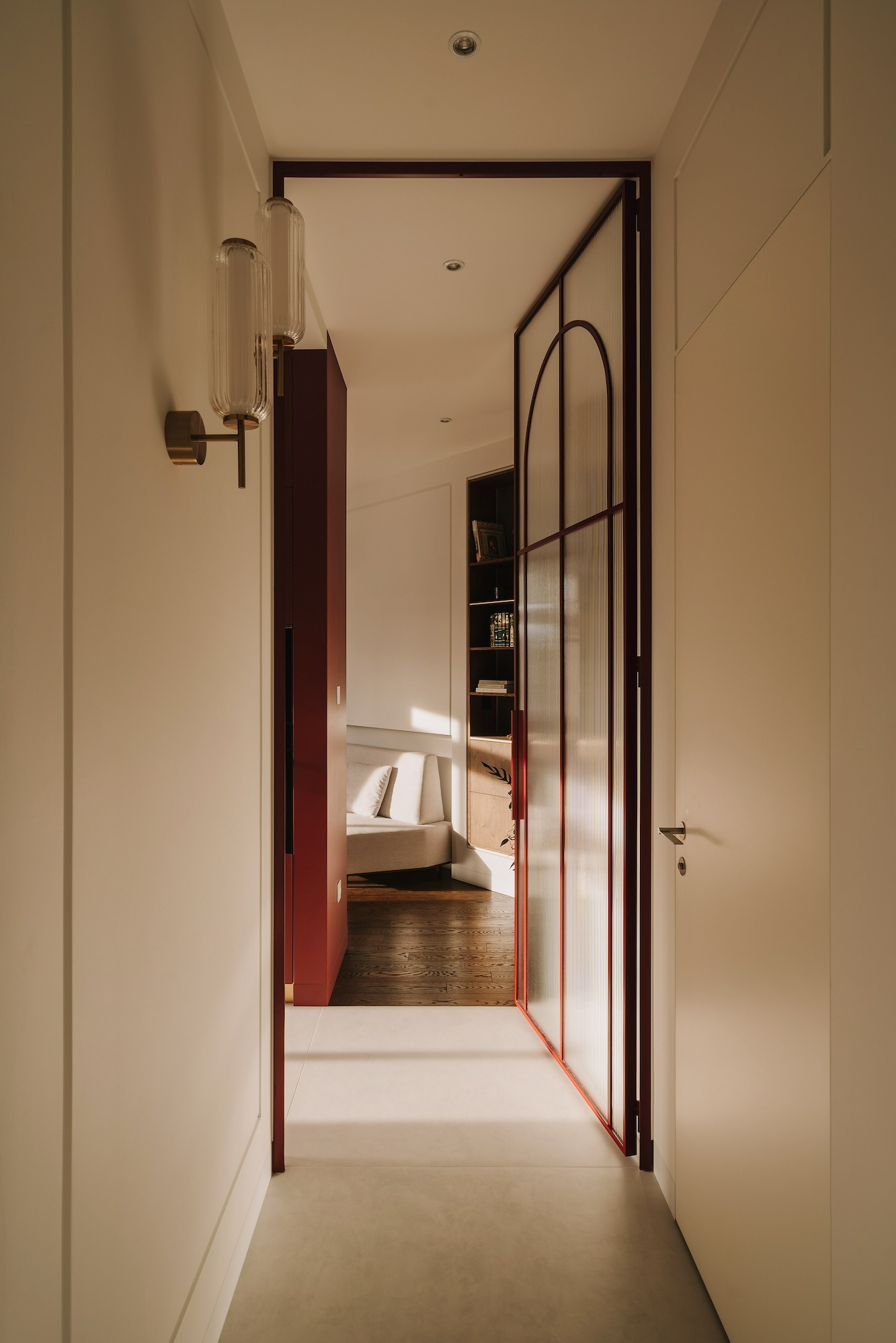 Wnętrze mieszkania z widokiem na salon przez przeszklone drzwi z czerwonymi ramami, oświetlone kinkietami i naturalnym światłem.