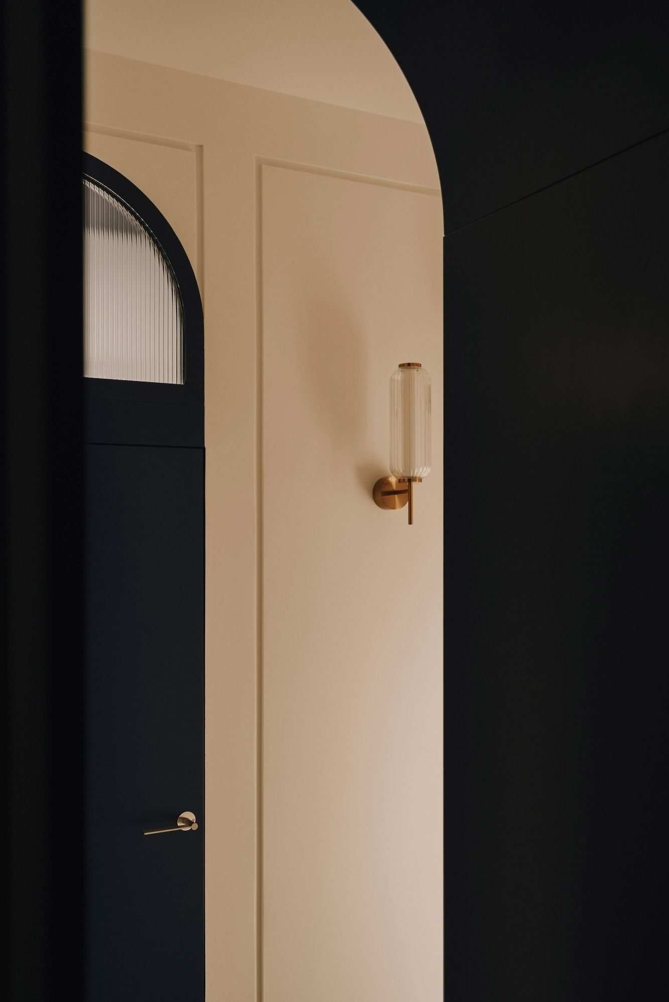 Częściowy widok wejścia z łukowym otworem, ciemnoniebieskie drzwi z ryflowanym szkłem i złotą klamką, beżowa ściana z panelem i szklanym kinkietem na złotym wsporniku.