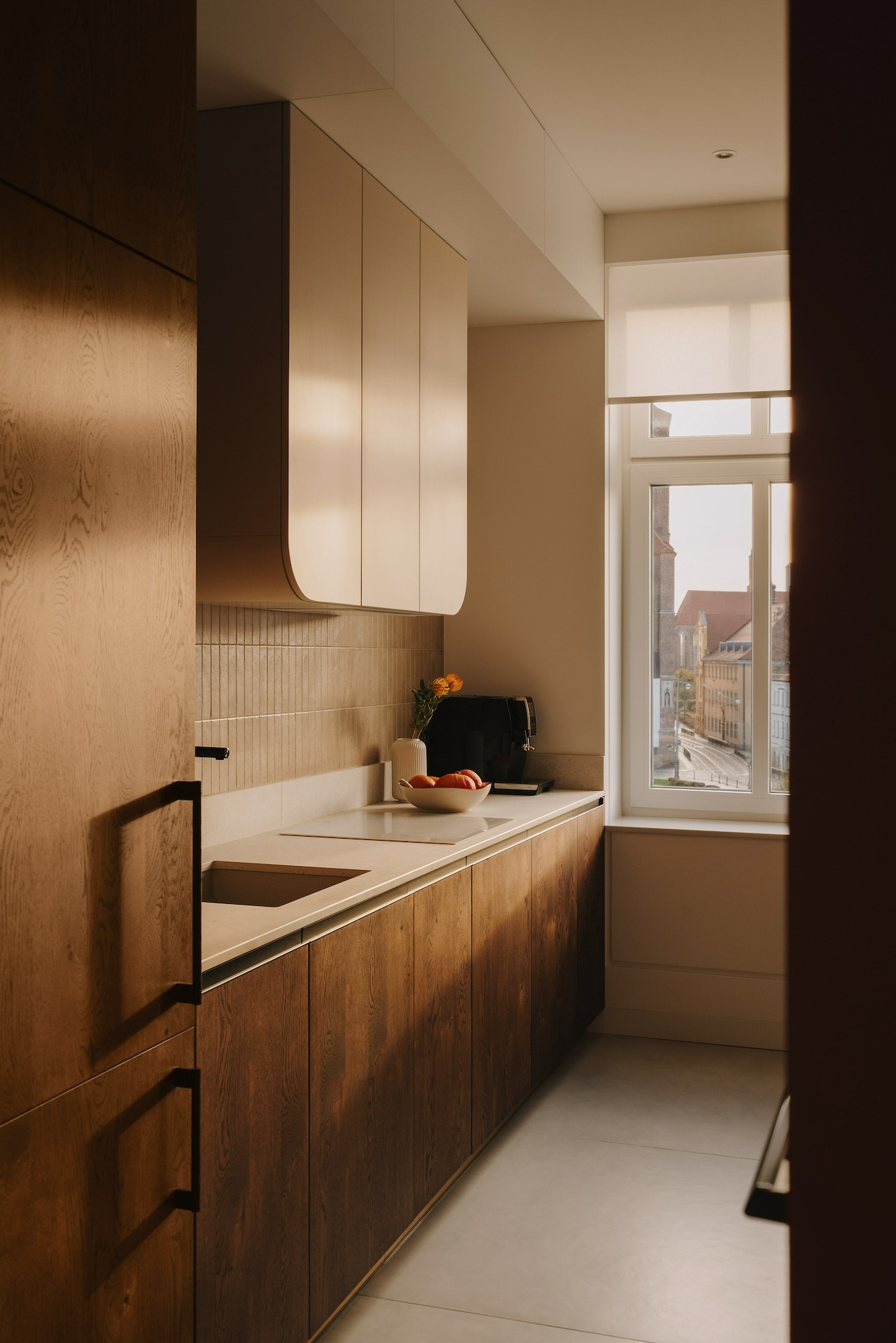 Nowoczesna kuchnia z drewnianymi szafkami w ciepłym odcieniu, jasnym blatem i widokiem na miasto za oknem, oświetlona popołudniowym słońcem.