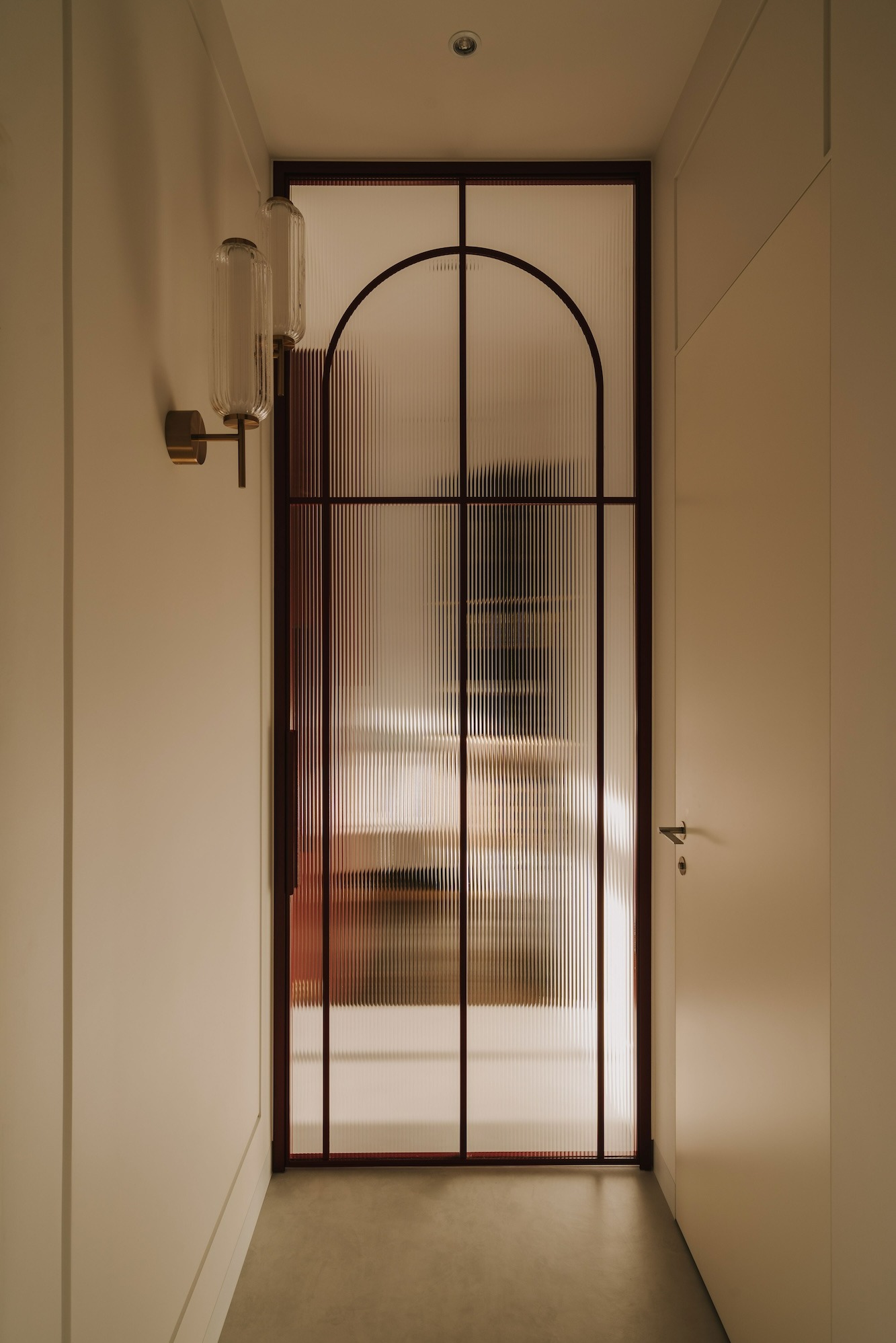 Korytarz z drzwiami z ryflowanego szkła w bordowej ramie i dwoma szklanymi kinkietami na ścianie.