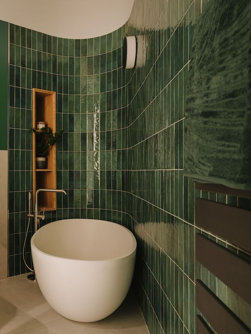 Nowoczesna łazienka z wolnostojącą, owalną wanną, zielonymi, pionowo ułożonymi płytkami z połyskiem, drewnianą półką z rośliną i grzejnikiem drabinkowym w kolorze ciemnego brązu.