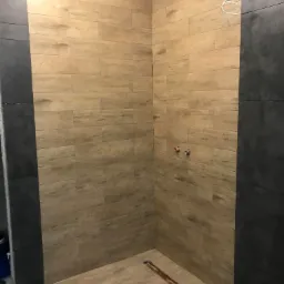 Wnętrze kabiny prysznicowej w trakcie wykańczania, z drewnopodobnymi płytkami na ścianach i podłodze, ciemnoszare płytki na ścianie bocznej, widoczne instalacje wodne i narzędzia glazurnicze.