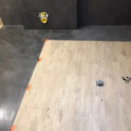 Fragment łazienki w trakcie prac wykończeniowych: częściowo położone ciemne płytki na ścianie i podłodze, jasna drewnopodobna podłoga z widocznymi otworami na instalacje, pomarańczowe kliny...