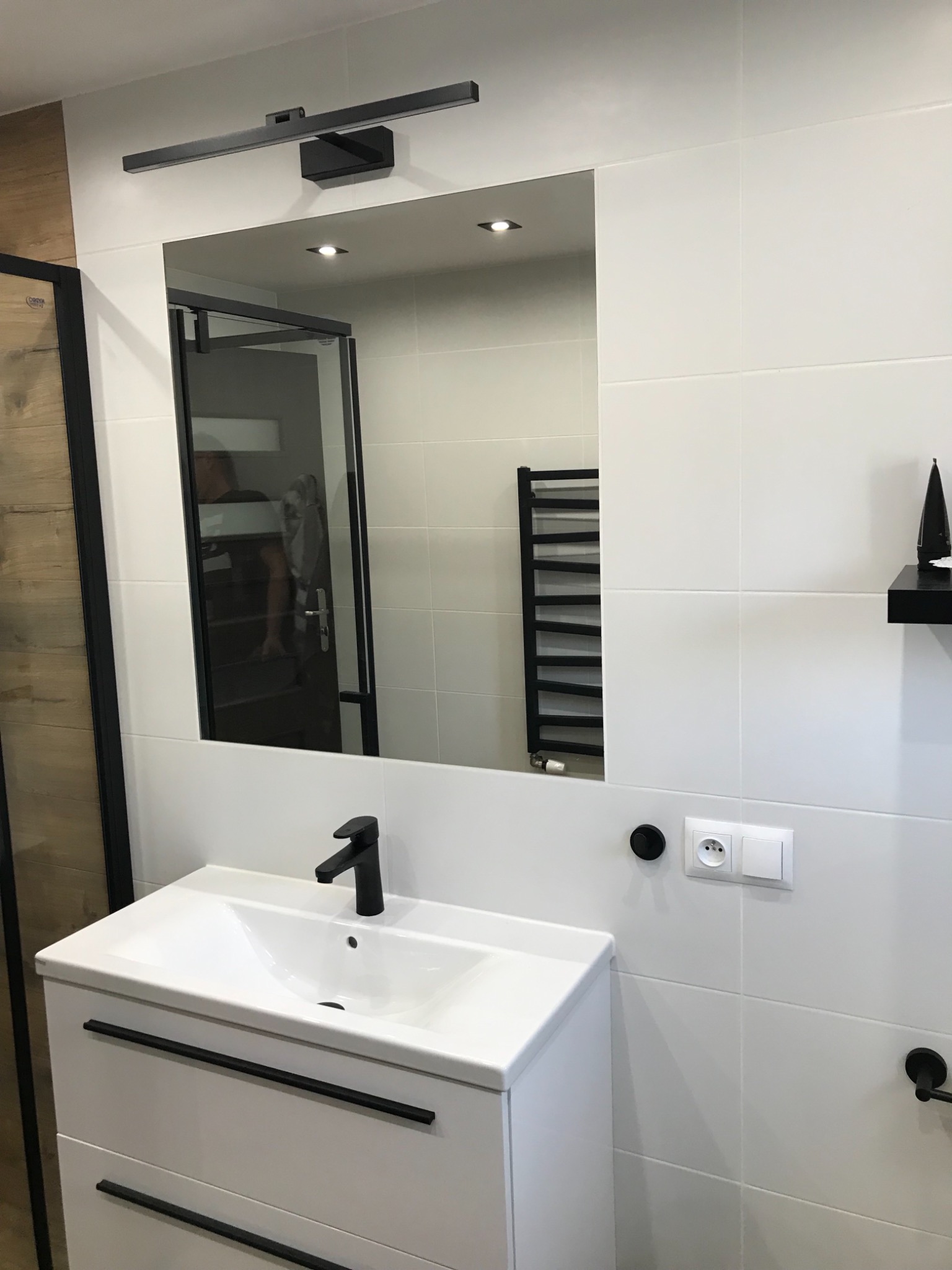Nowoczesna łazienka z białymi płytkami, czarną armaturą i oświetleniem, umywalka z szafką, lustro z podświetleniem LED, widoczny fragment kabiny prysznicowej.