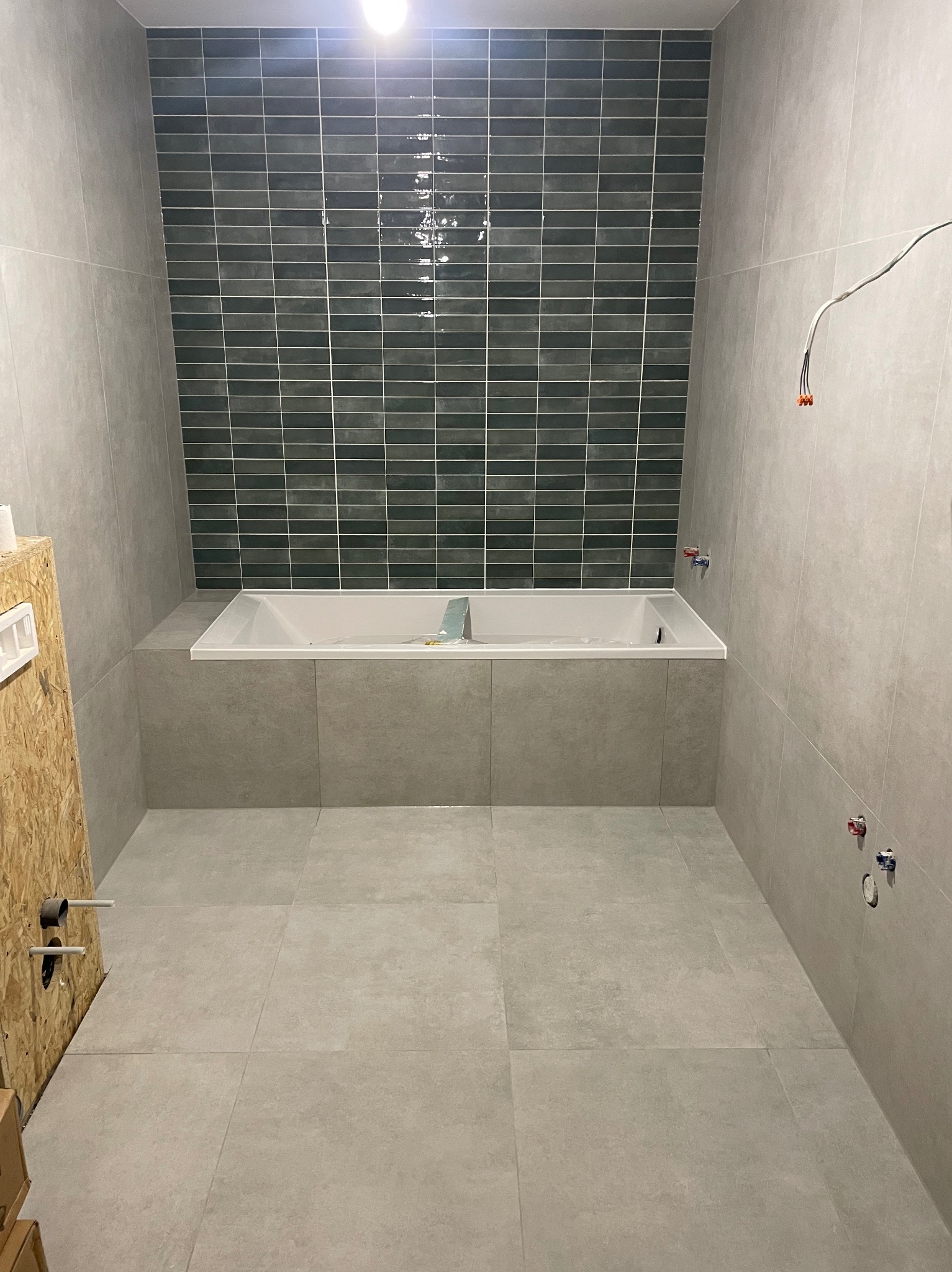 Nowoczesna łazienka w szarościach z wanną obudowaną płytkami i akcentem ciemnozielonej mozaiki na ścianie. Widoczne przyłącza wodne i elektryczne.