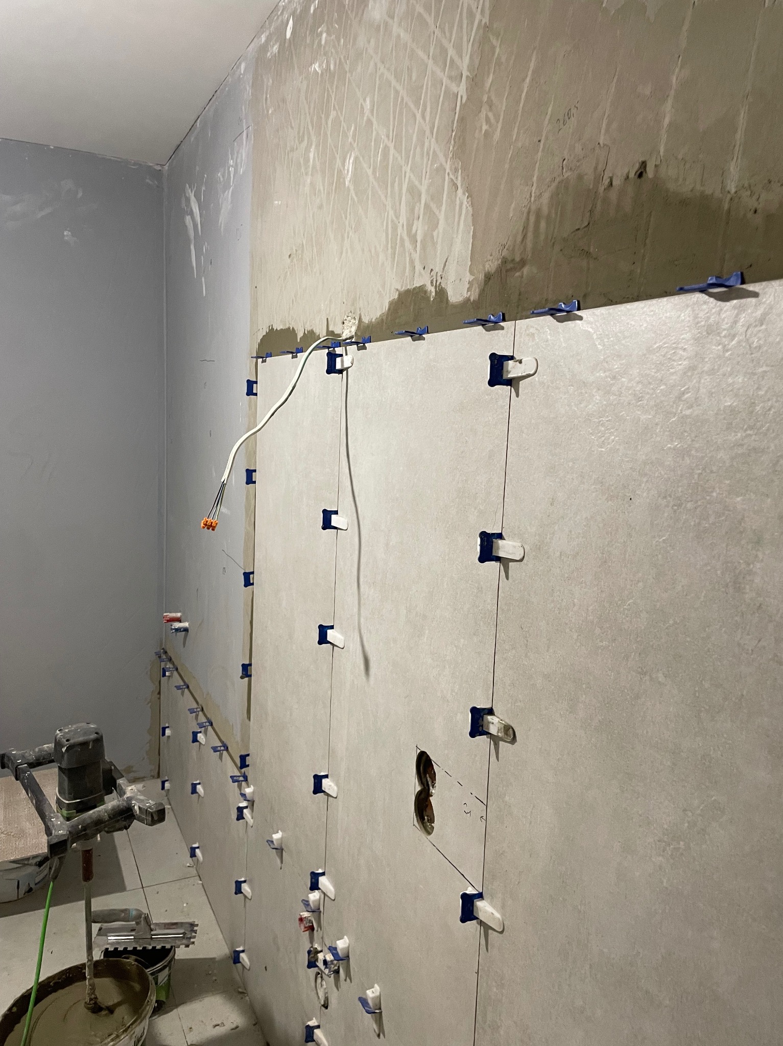 Ściana w trakcie układania płytek z użyciem klipsów poziomujących. Widoczny mikser do zaprawy i przygotowana powierzchnia ściany.