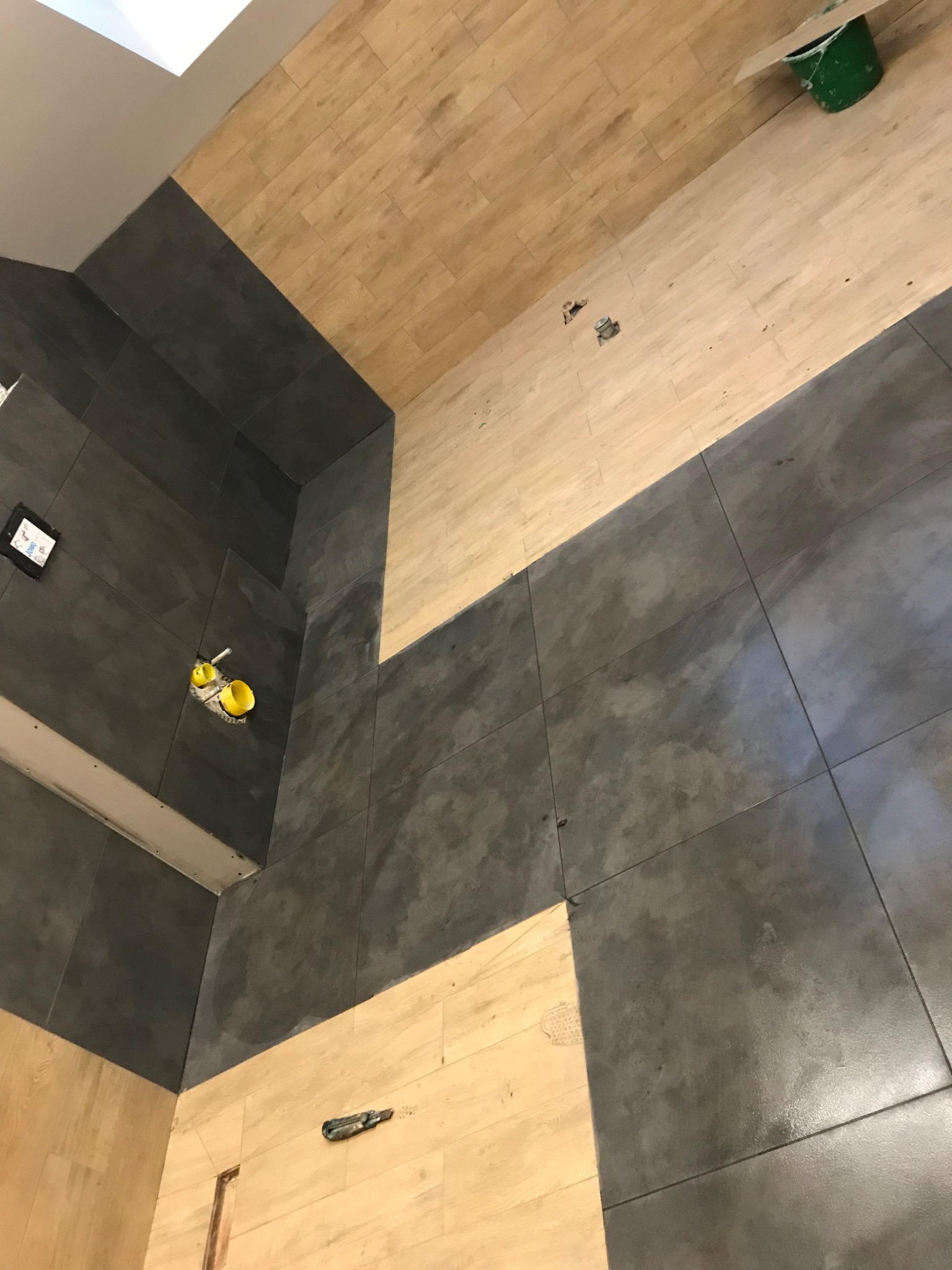 Fragment łazienki w trakcie wykańczania, widoczne szare płytki na ścianach i podłodze, oraz jasne panele na ścianach. W tle widoczne żółte osłonki na rury i zielone wiadro.