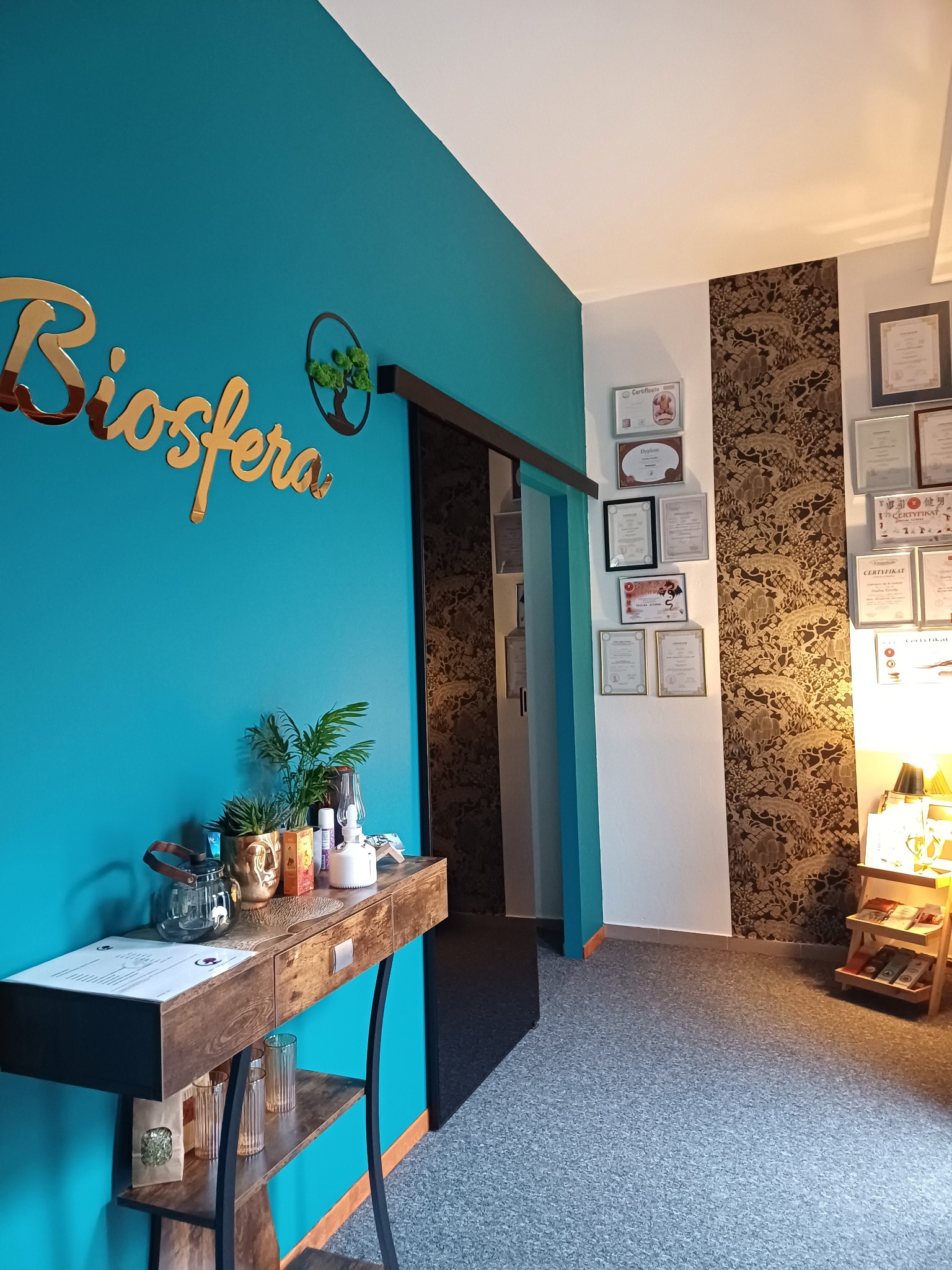 Wnętrze gabinetu z turkusową ścianą i złotym napisem 'Biosfera', drewniany stolik z dekoracjami roślinnymi i szklanym czajnikiem, na ścianie certyfikaty i dyplomy, wzorzysta tapeta i szara podłoga.