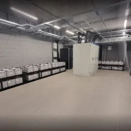 Wnętrze pomieszczenia technicznego z rzędami akumulatorów na metalowych regałach, szafą sterowniczą i widocznymi przewodami pod sufitem, oświetlone lampami jarzeniowymi.