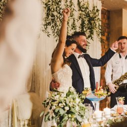 Gie Studio - Uśmiechnięta para nowożeńców wznosi ręce w geście triumfu podczas wesela, w tle dekoracje z zielonych roślin i biała zasłona, obok muzycy z saksofonem, na pierwszym planie stół z kwiatami i drinkami.