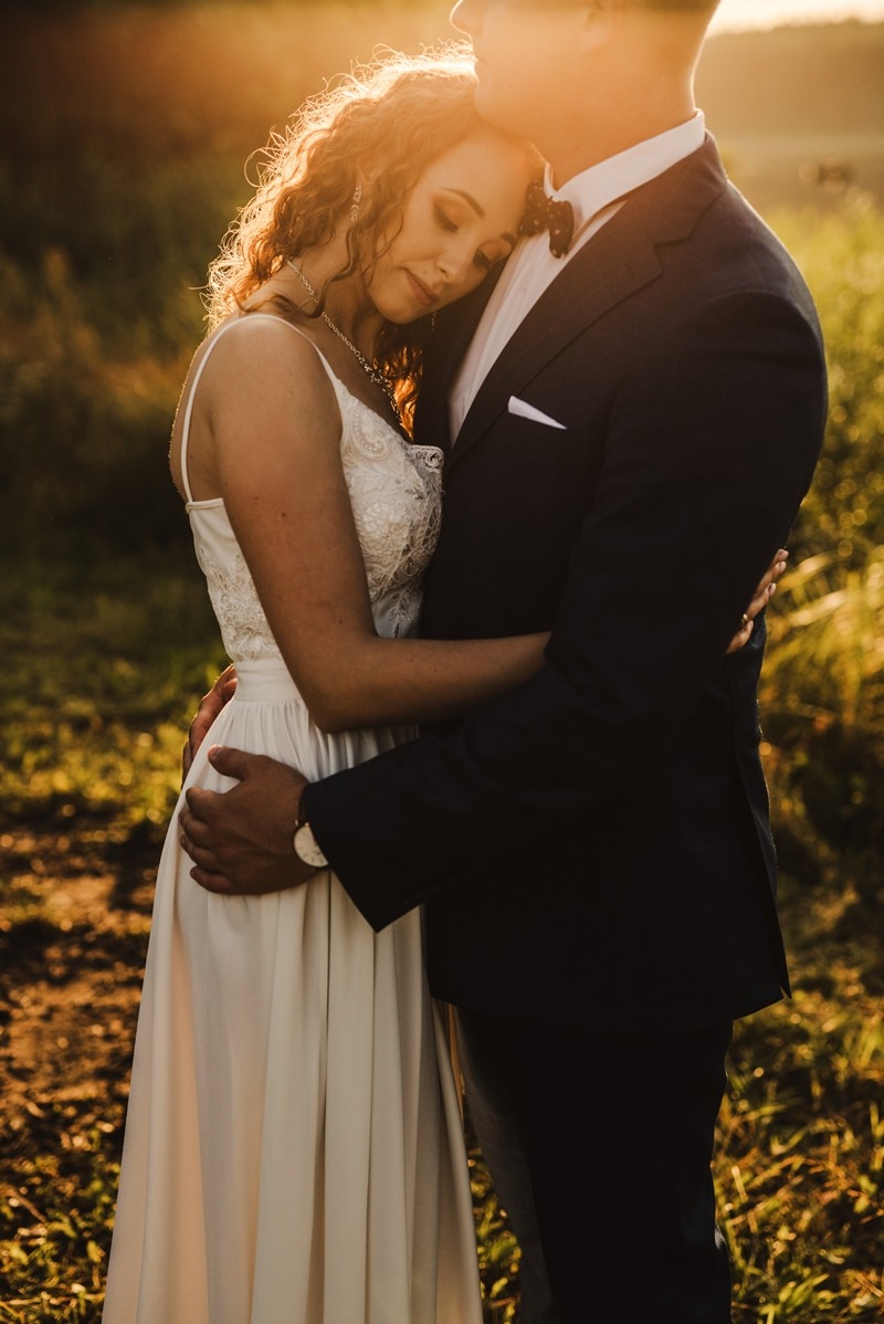 Para młoda w objęciach na tle zachodzącego słońca, panna młoda z kręconymi włosami opiera głowę na ramieniu pana młodego w granatowym garniturze.