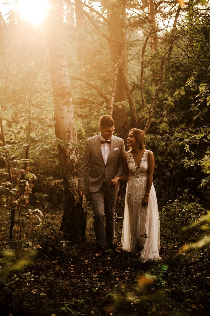 Para młoda spaceruje po lesie trzymając się za ręce, w tle prześwitujące promienie słoneczne.