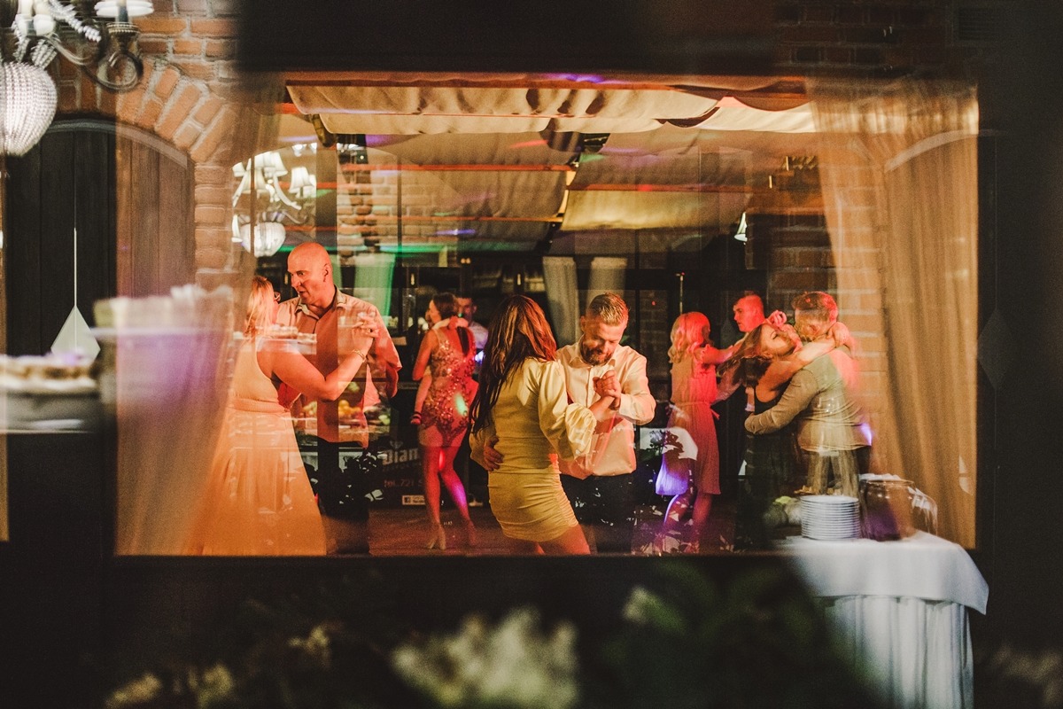 Grupa osób tańczących na parkiecie oświetlonym kolorowymi światłami, widoczna przez okno z refleksami, w tle ceglana ściana i dekoracje.