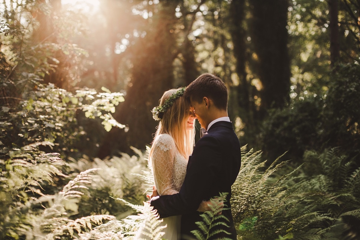 Para młoda w objęciach w lesie, panna młoda w koronkowej sukni i wianku, delikatne promienie słoneczne przebijają się przez drzewa.