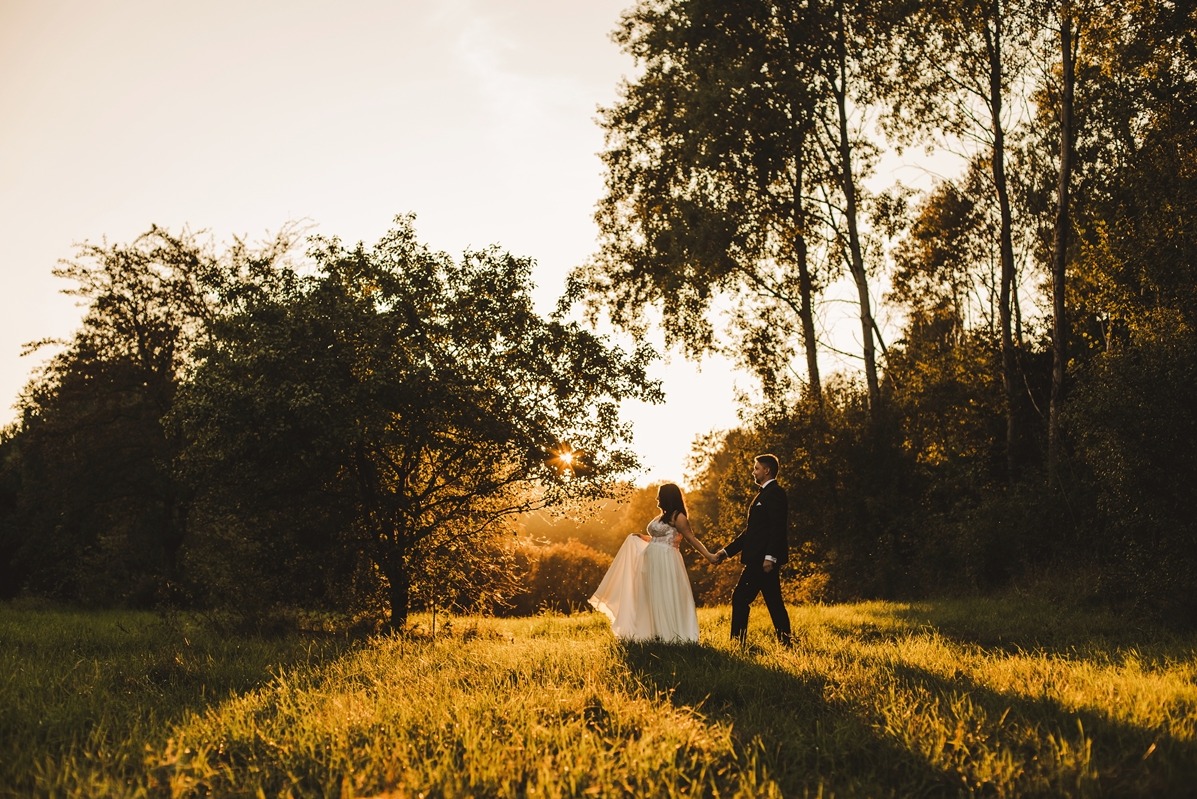 Para młoda trzymająca się za ręce spaceruje po łące oświetlonej zachodzącym słońcem, widoczne długie cienie traw i drzew.