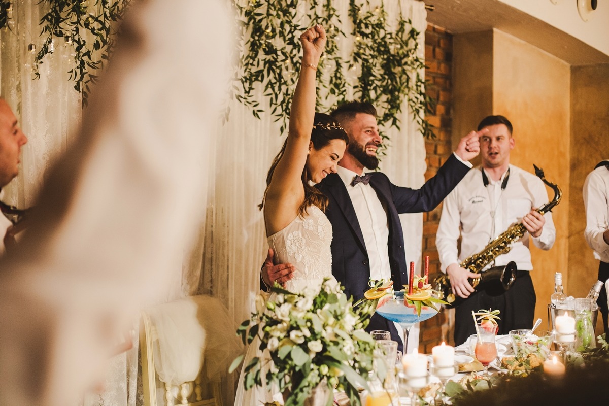 Uśmiechnięta para nowożeńców wznosi ręce w geście triumfu podczas wesela, w tle dekoracje z zielonych roślin i biała zasłona, obok muzycy z saksofonem, na pierwszym planie stół z kwiatami i drinkami.