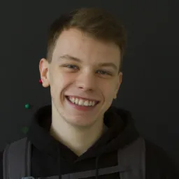 Portret uśmiechniętego, młodego mężczyzny z plecakiem na tle ciemnej ściany z pinezkami, Siemianowice Śląskie.