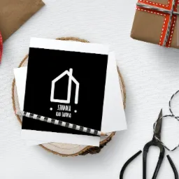 Karta z minimalistycznym logo domu 'Stodoła nad Tanwią' na czarnym tle, ułożona na drewnianym plastrze, obok ołówek w czarno-białe paski, prezent zapakowany w brązowy papier, sznurek i nożyczki...