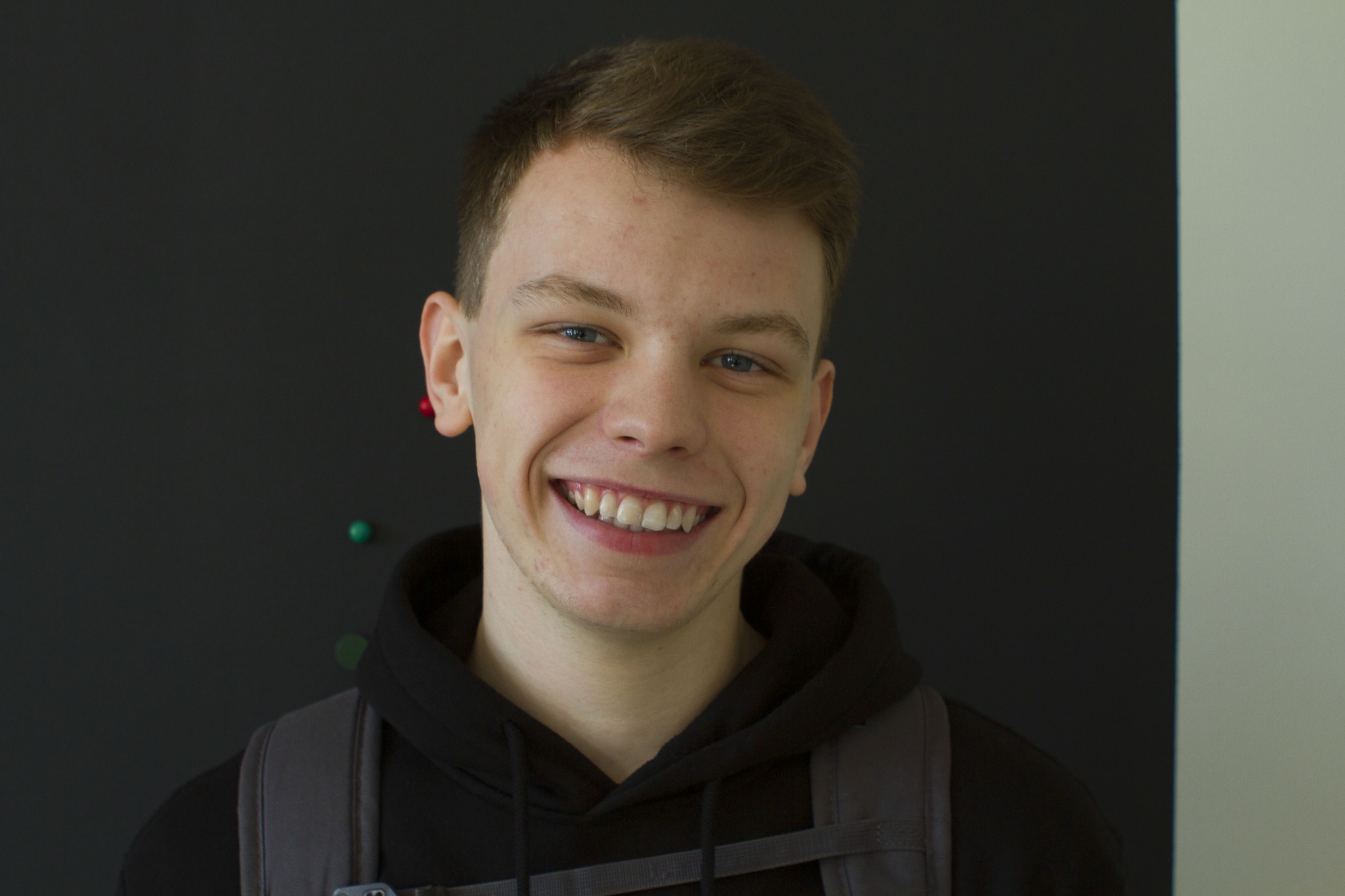 Portret uśmiechniętego, młodego mężczyzny z plecakiem na tle ciemnej ściany z pinezkami, Siemianowice Śląskie.