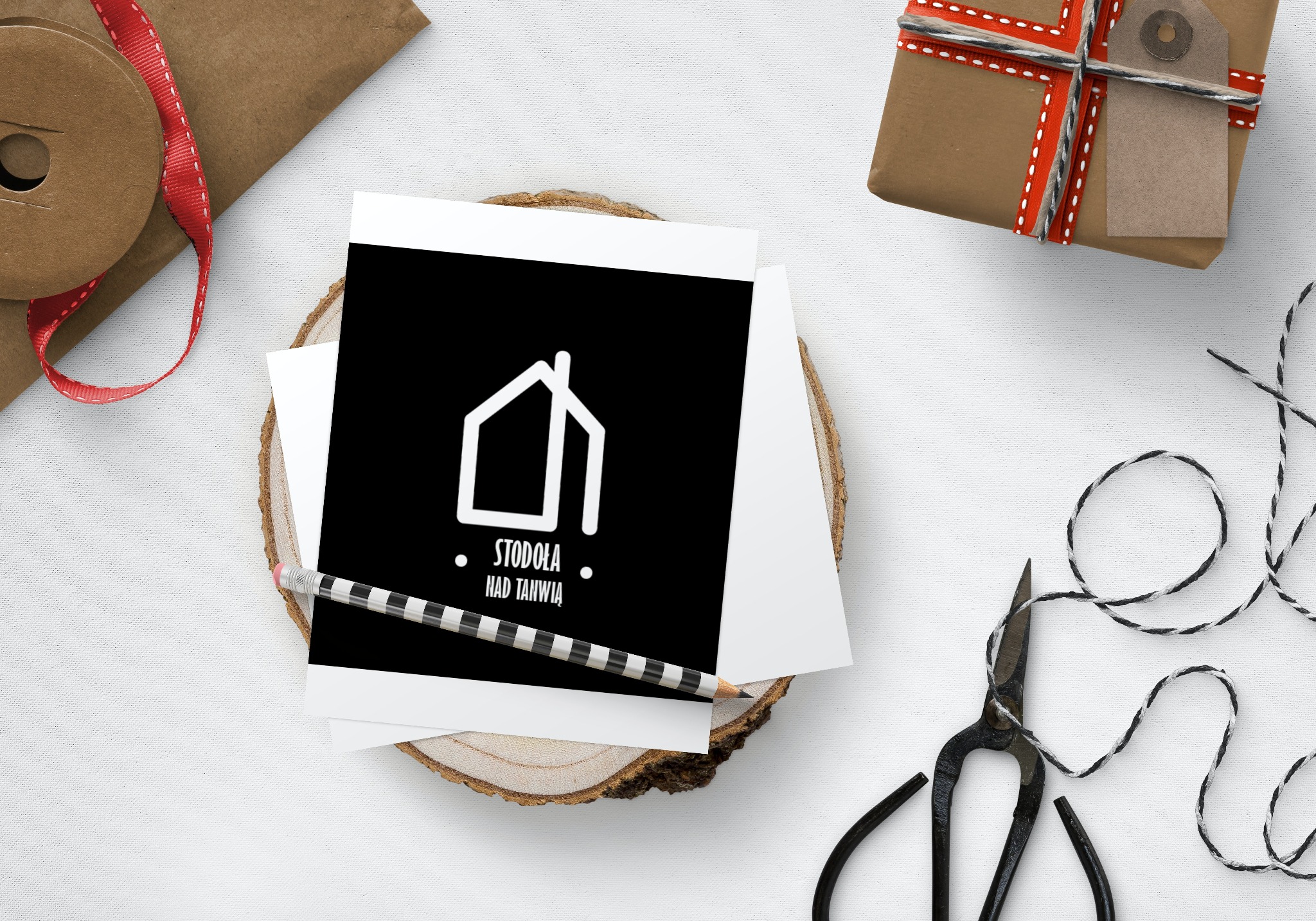 Karta z minimalistycznym logo domu 'Stodoła nad Tanwią' na czarnym tle, ułożona na drewnianym plastrze, obok ołówek w czarno-białe paski, prezent zapakowany w brązowy papier, sznurek i nożyczki...