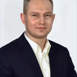 Portret mężczyzny w ciemnym garniturze i białej koszuli na jasnym tle, sugerujący profesjonalizm i zaufanie.