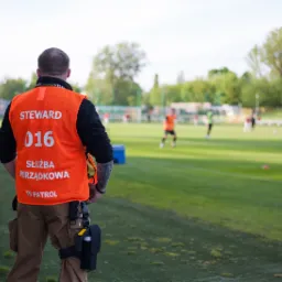 Ochroniarz w pomarańczowej kamizelce z napisem 'STEWARD' i numerem 016, stojący tyłem na tle boiska piłkarskiego z zawodnikami.