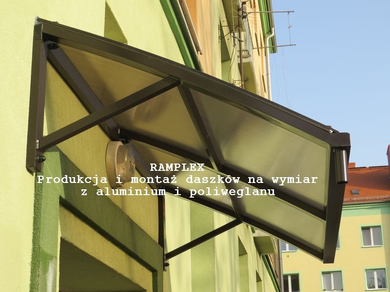 Brązowy daszek z poliwęglanu zamontowany nad wejściem do budynku, widoczne mocowania do ściany i elementy konstrukcyjne.