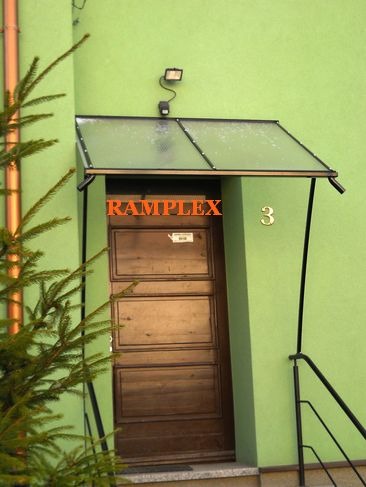 Wejście do budynku z drewnianymi drzwiami, nad którymi zamontowano prostą, przezroczystą osłonę z czarną ramą i wspornikami, oświetlone lampą nad osłoną, z numerem 3 na ścianie.