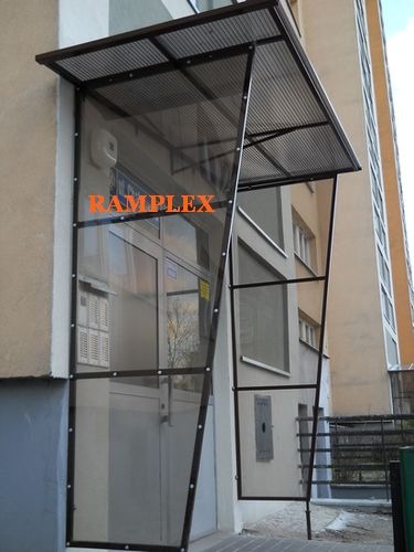 Zadaszenie wejścia do budynku z przezroczystym wypełnieniem i metalową konstrukcją, widoczne skrzynki pocztowe i instalacje na ścianie.