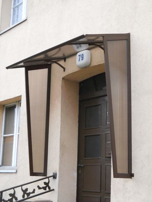 Brązowy daszek z bocznymi osłonami nad wejściem do budynku z numerem 79, widoczne schody z metalową balustradą i okno z żaluzjami.