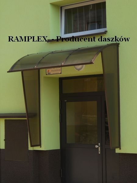 Zadaszenie nad wejściem do budynku, wykonane z przezroczystego tworzywa sztucznego z brązowymi ramami, widoczne logo producenta na elewacji budynku.