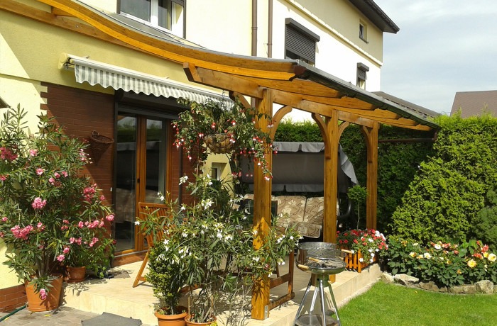 Zadaszona drewniana konstrukcja z łukowatymi belkami, wiszące donice z kwiatami, grill na tarasie i huśtawka w tle.