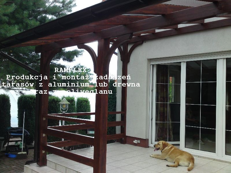 Zadaszony taras z drewnianą konstrukcją, widoczne elementy łukowe i brązowe belki, na podłodze leży pies rasy labrador, w tle krzewy i okno przesuwne.