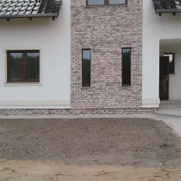 Nowy dom jednorodzinny z jasną elewacją, fragment ściany wykończony cegłą klinkierową, widoczne okna z brązowymi ramami oraz fragment dachu z ciemną dachówką, teren przed budynkiem jeszcze...