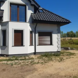 Nowoczesny dom jednorodzinny z białymi ścianami, brązowymi roletami i ciemną dachówką, fragment elewacji z oknami i widoczną instalacją fotowoltaiczną na dachu.