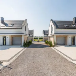 Nowoczesne szeregówki z jasną elewacją i grafitowym dachem, wykończone kostką brukową i żwirową drogą dojazdową, widok z perspektywy wjazdu.