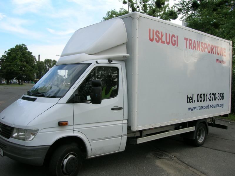 Biały samochód dostawczy z nadrukiem 'Usługi Transportowe Wrocław' oraz numerem telefonu i adresem strony internetowej, zaparkowany na ulicy.