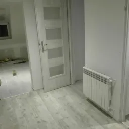 Wnętrze po odświeżeniu: białe ściany, szare panele podłogowe imitujące drewno, biały grzejnik i częściowo otwarte białe drzwi z przeszkleniami, widoczne narzędzia w sąsiednim pomieszczeniu.