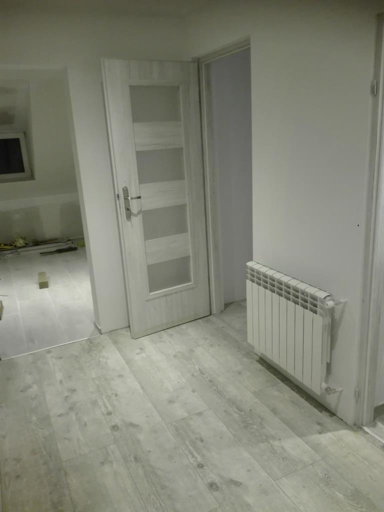 Wnętrze po odświeżeniu: białe ściany, szare panele podłogowe imitujące drewno, biały grzejnik i częściowo otwarte białe drzwi z przeszkleniami, widoczne narzędzia w sąsiednim pomieszczeniu.